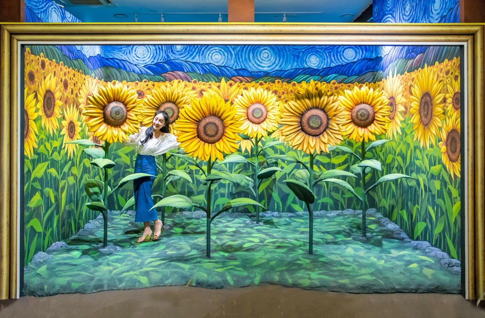 Girl smiling for a photo with a sunflower art piece inside Alive Museum