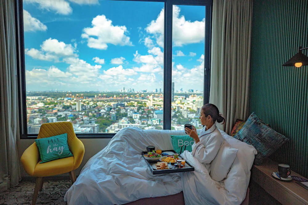 Woman enjoying breakfast in bed with panoramic Bangkok skyline view at AVANI Sukhumvit