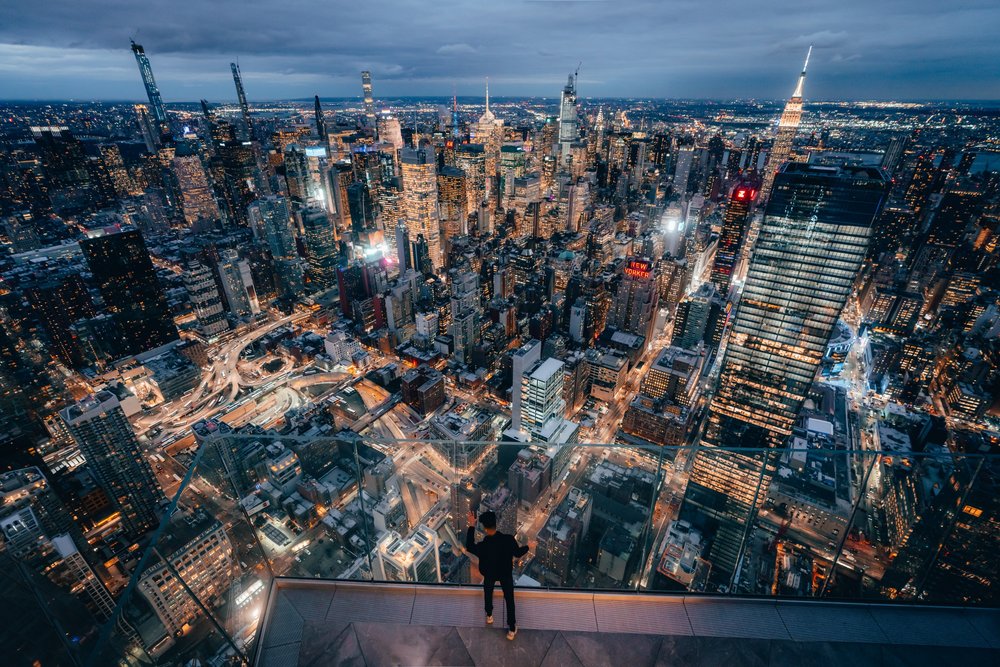 Person standing on The Edge observation deck at night overlooking New York City skyline.