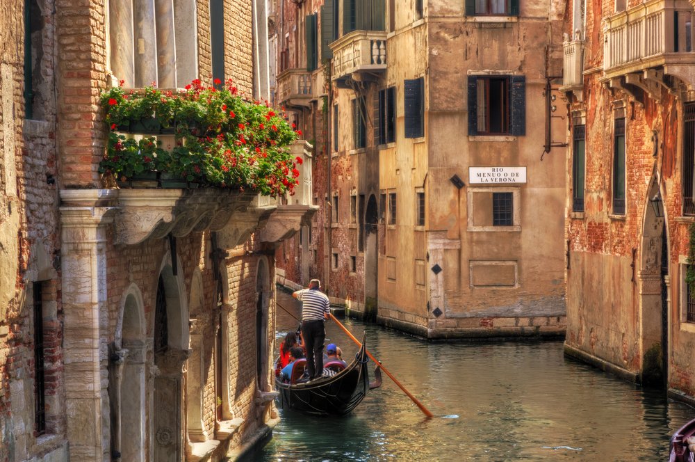Gondola with passengers cruising down narrow Venetian canal beside flowered balconies