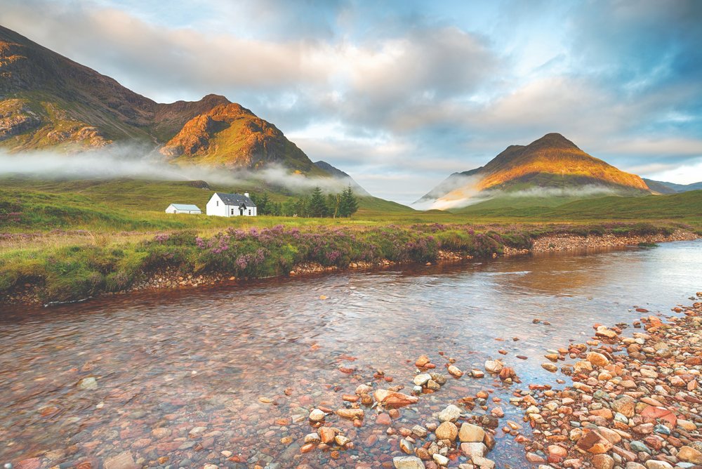 White cottage in Glencoe with misty mountains and sunrise lighting the Highlands