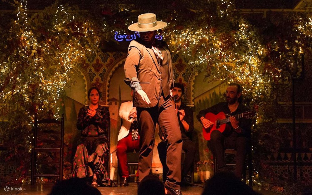 A man performs in the middle of the stage, while behind him sit the guitarist, the singer, and the palmeros (clappers)