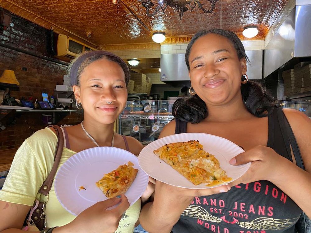 Two friends smiling and holding pizza slices inside a NYC pizzeria
