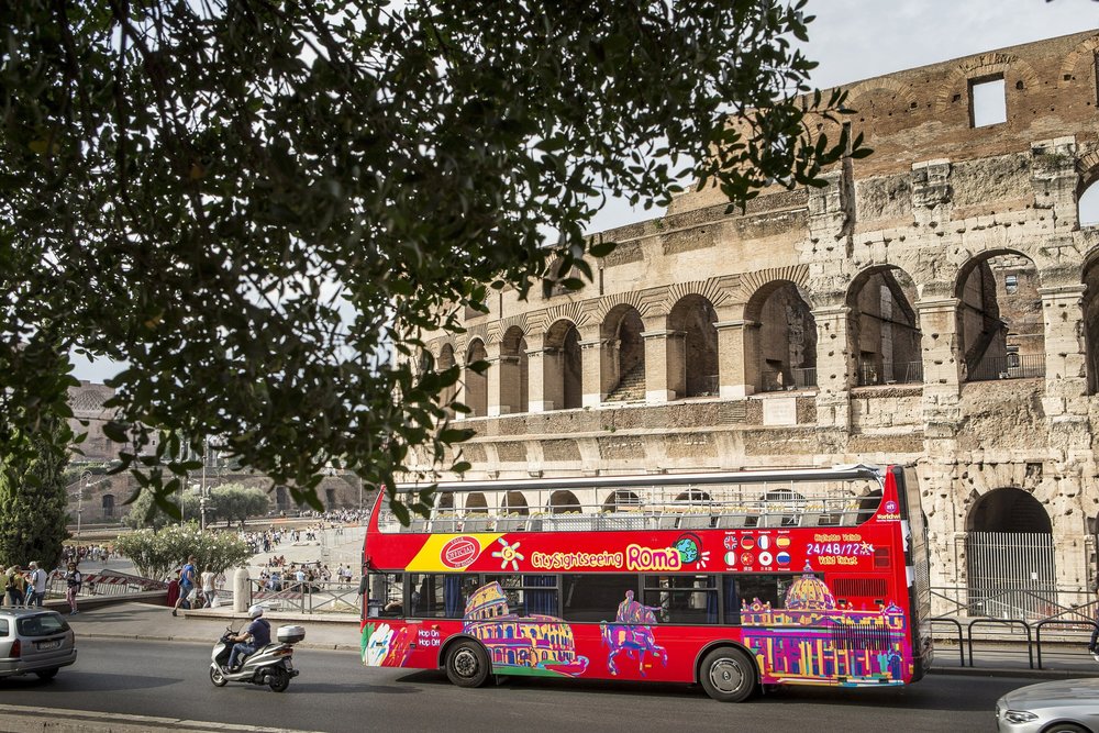 Red hop on hop off bus driving beside the Colosseum in Rome
