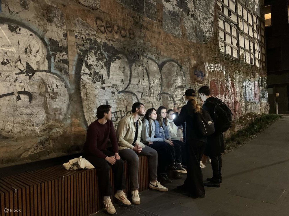In an alley, a group of tourists are sitting down, while their two guides stand