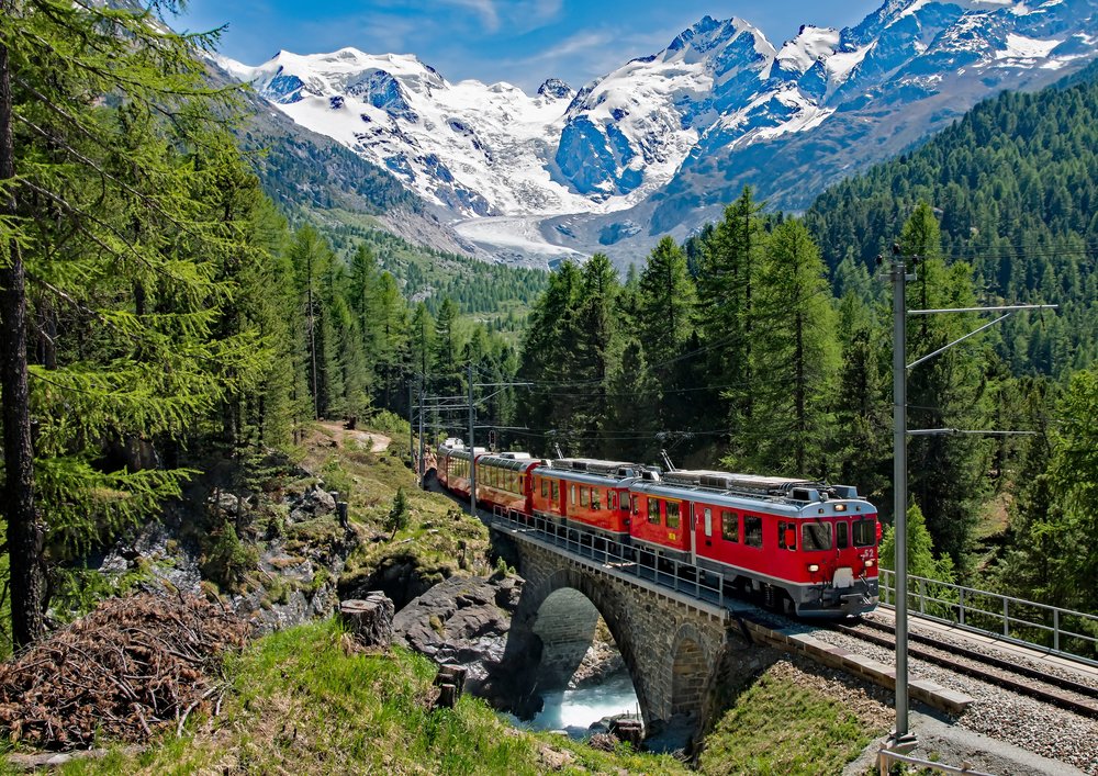 Red Bernina Express train traveling through forests toward snowy mountains
