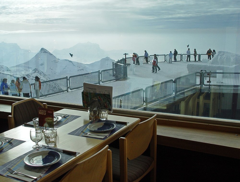 A table beside a glass window inside the Piz Gloria. Outside, people are walking on snow.