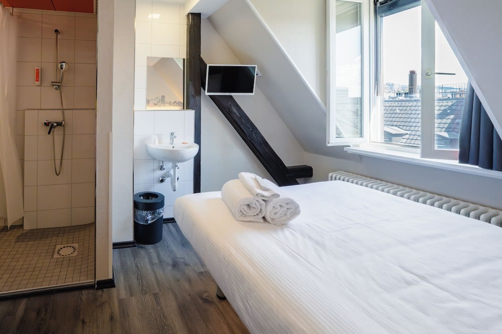 A bed is pushed to the side, where a window overlooks the city. To the bed’s left is a small bathroom
