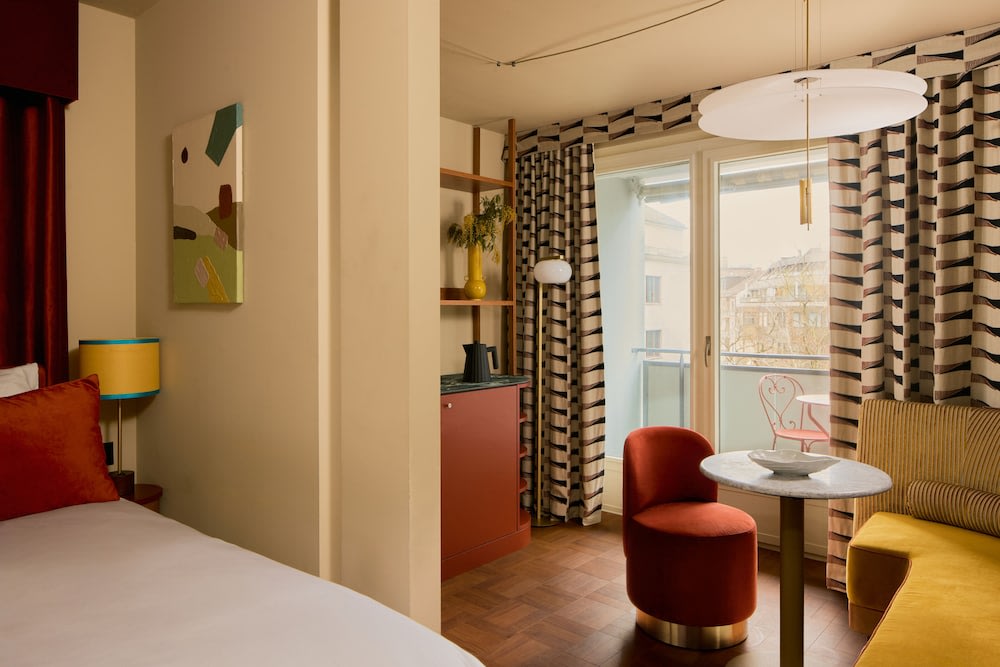 The hotel room, which shows a portion of the bed, a small living area, and a balcony