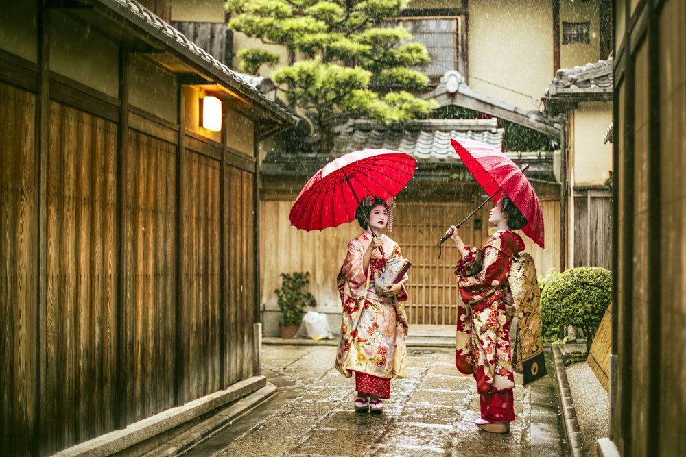 Two maiko holding red umbrellas in a rainy wooden alley in Gion, Kyoto