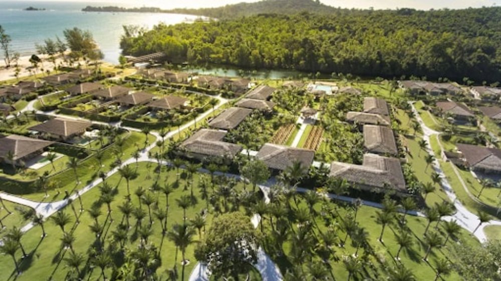 Fusion Resort Phu Quoc from above. There are many palm trees and vegetation surrounding the halls and accommodation. A view of a river connecting to the sea is also seen.