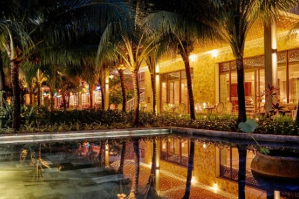 A picture of Salinda Resort Phu Quoc Island’s pool can be seen showered by the warm lights of the building next to it. There are many plants and palm trees surrounding the water.