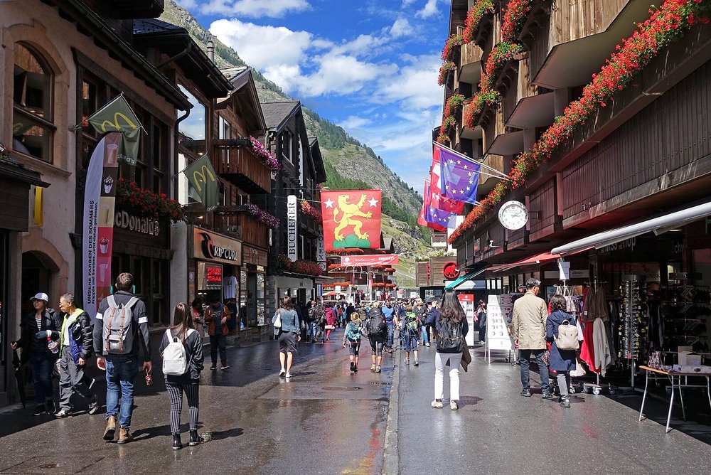 Crowded Swiss alpine village street with shops, flags, and mountain scenery