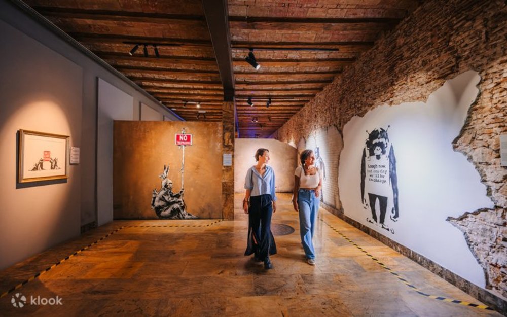 Visitors walking through an indoor exhibition space featuring large Banksy-style wall artworks.