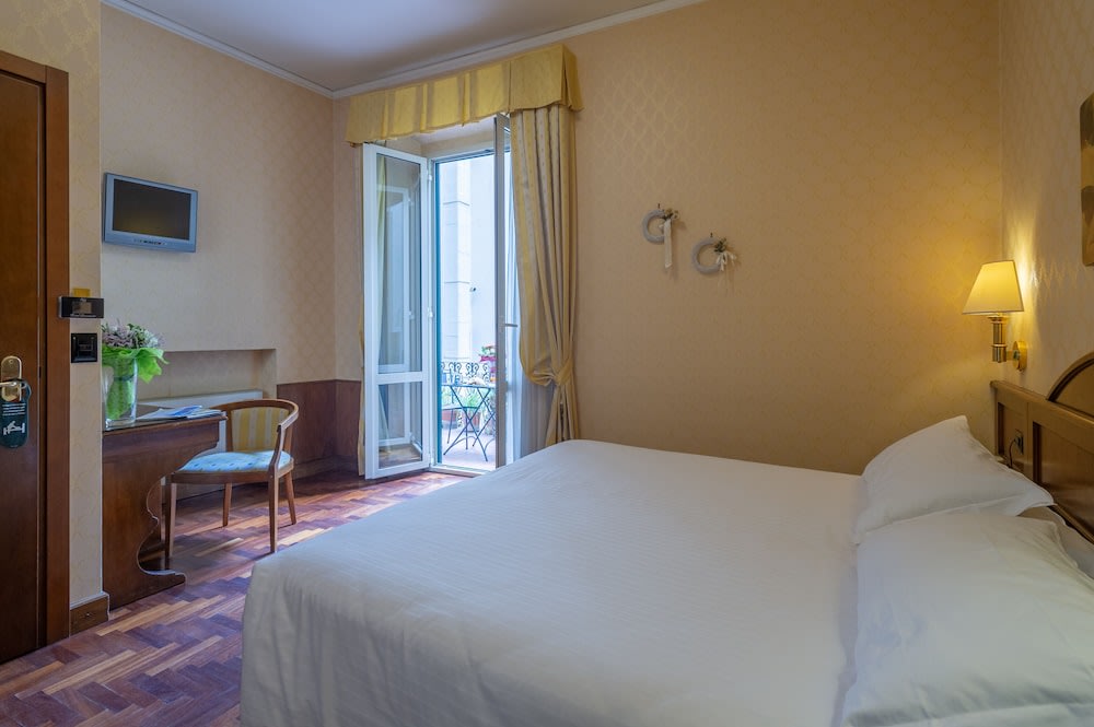 At the foot of a bed is a small desk and a chair, while the other side has an open window leading to a balcony