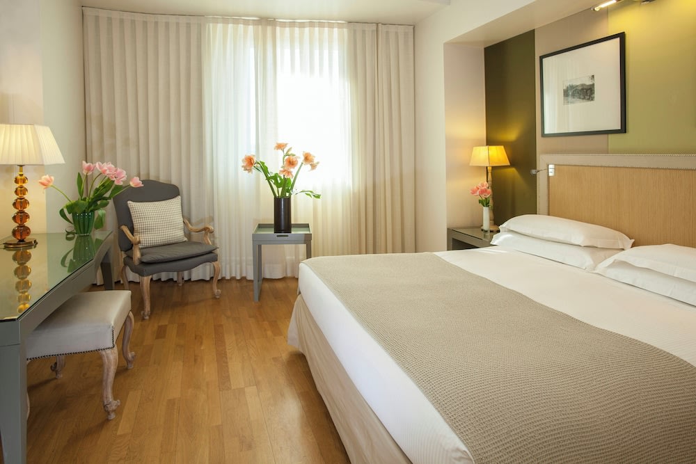 On the foot of the bed is a desk and a chair, while at its side is a chair and large window with curtains drawn shut