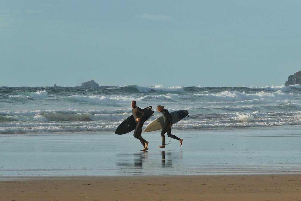 Surfers carrying boards while walking along a windy beach coastline | Photo Credit: Belinda Fewings on Unsplash