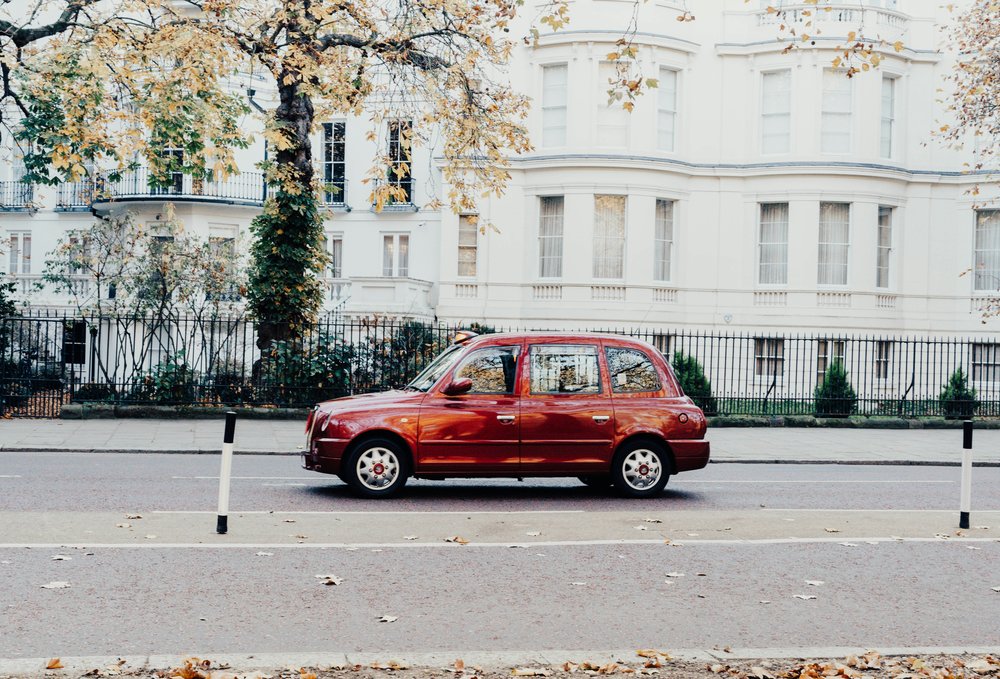 Red car driving past white townhouses on a quiet street in London | Photo Credit: Anna Claire Schellenberg on Unsplash