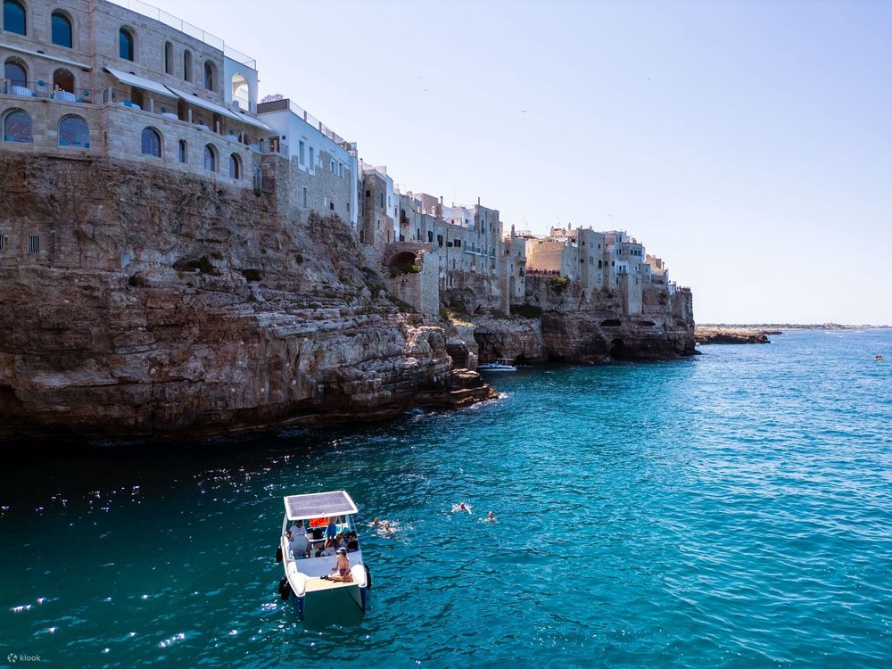 Short buildings on top of a large cliff structure beside a body of water with a floating white boat