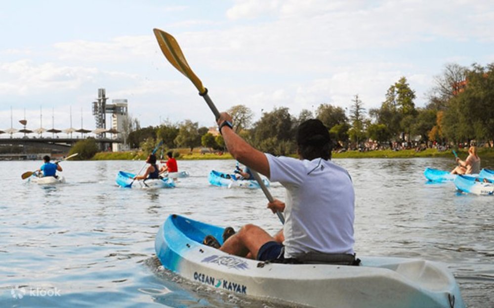 People kayaking on the Guadalquivir River in Seville with trees and city structures in the background.