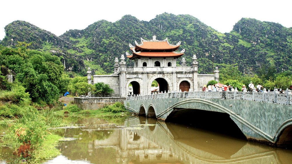 A traditional stone gate with a tiered red roof stands at the end of a bridge over a calm river in Ninh Binh