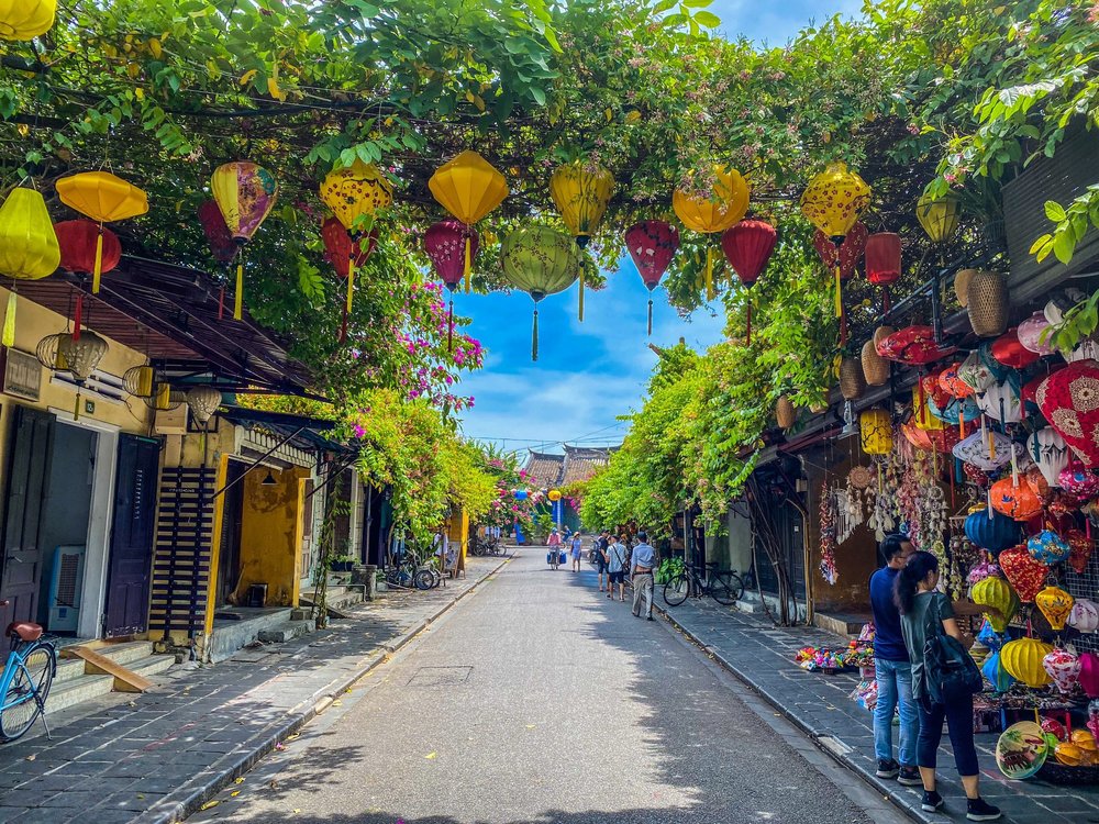 A narrow street in Hoi An lined with yellow buildings and overhead greenery, decorated with vibrant silk lantern