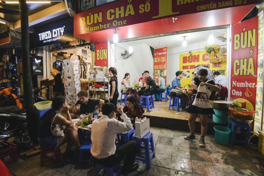 Travelers and locals sit on low plastic stools outside a brightly lit Bun Cha restaurant in a busy city at night
