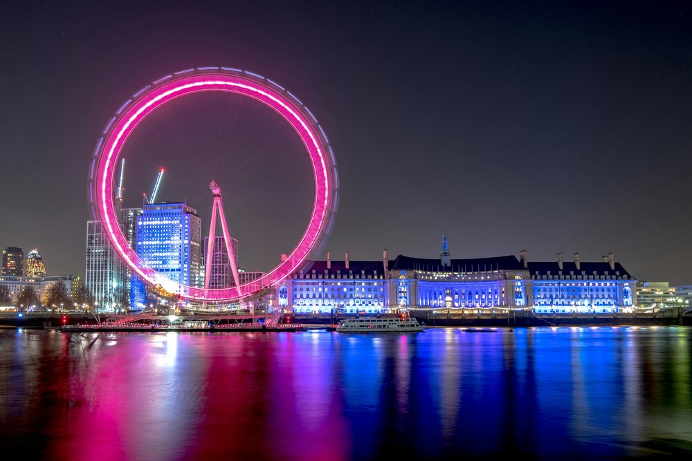 London Eye at night illuminated in pink with city lights reflecting on the Thames