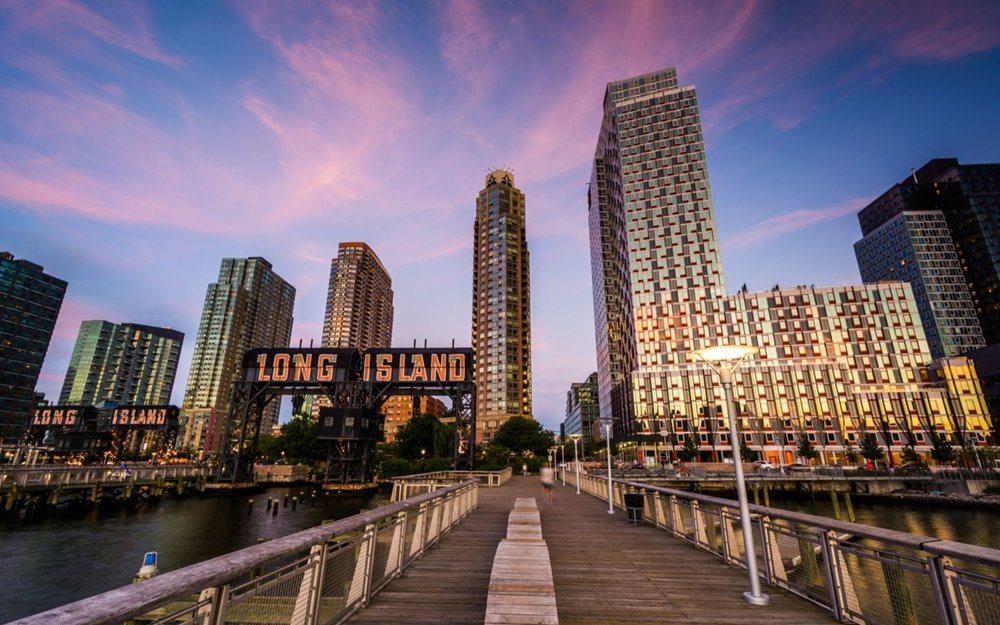 The point of view of a person walking on a pier toward tall buildings and a sign that reads “Long Island” against a sunset