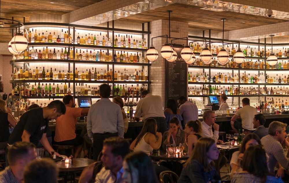 A crowded, dimly lit bar with a floor-to-ceiling display of drinks