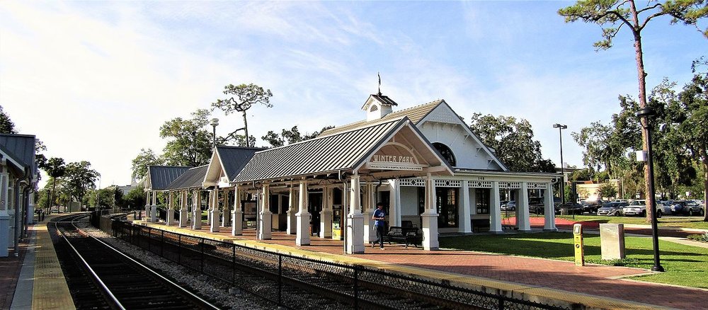 View of the Winter Park train station platform with tracks, a white station building, and trees in the background