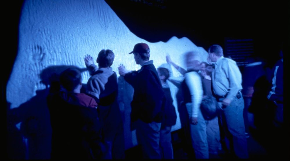Group of visitors placing hands on a large iceberg display inside Titanic: The Artifact Exhibition, lit in blue
