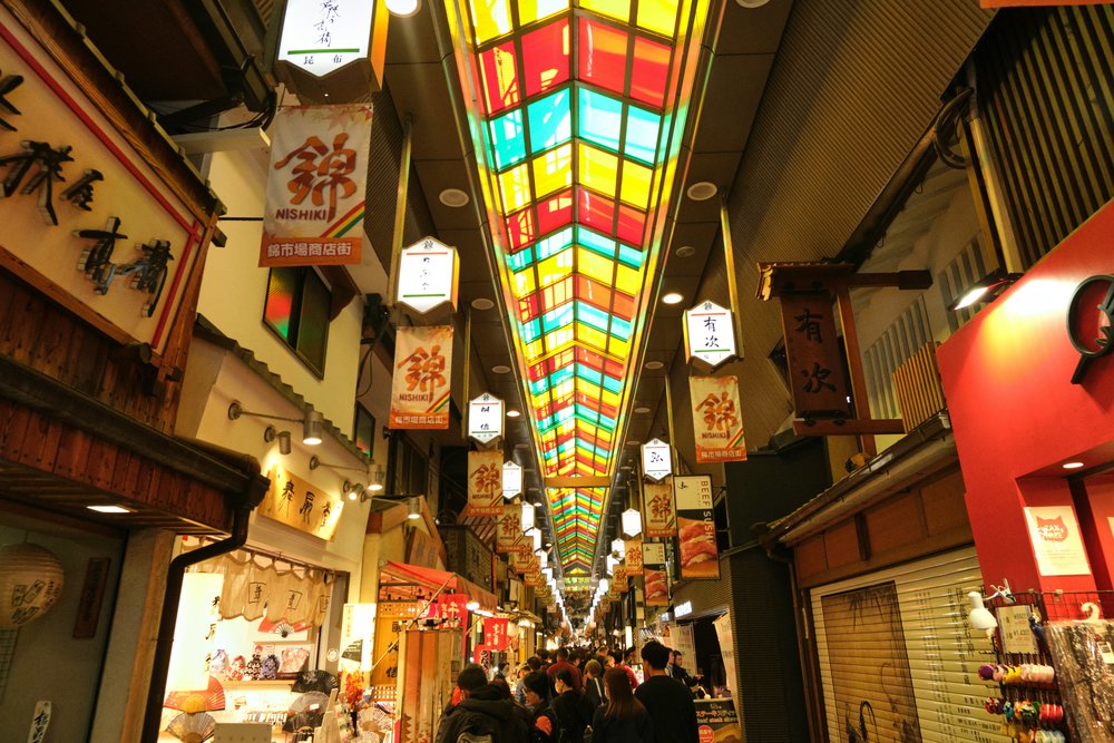 The colorful, bustling Nishiki Market in Kyoto, with shops selling various goods under bright, overhead lights | Photo Credit: Rebecca Clarke on Unsplash