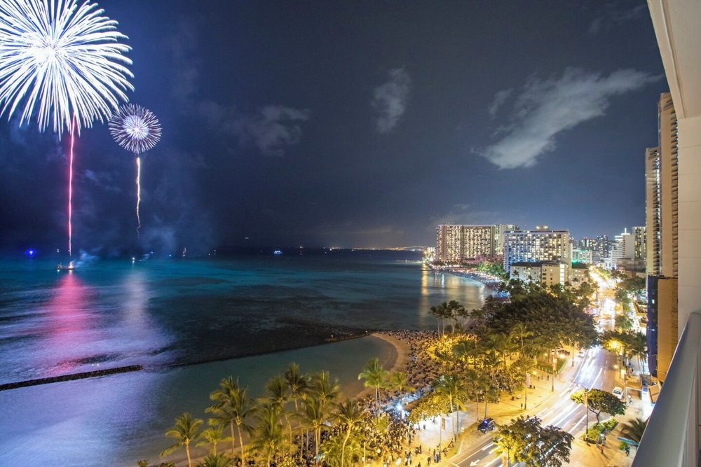 A high-angled view of a beach full of people and buildings below a night sky with large fireworks.