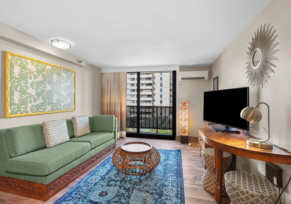 The interior of a colorful hotel room decorated with a couch, carpet, tables, a tv, lamps and other small furniture.