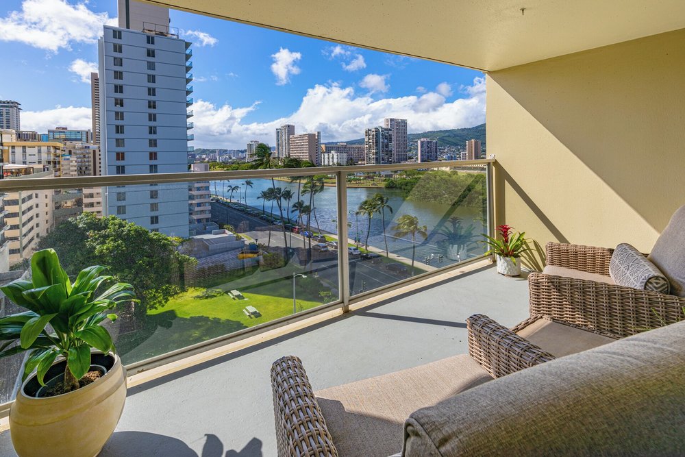 The view of buildings, a body of water, and greenery from a balcony with chairs.