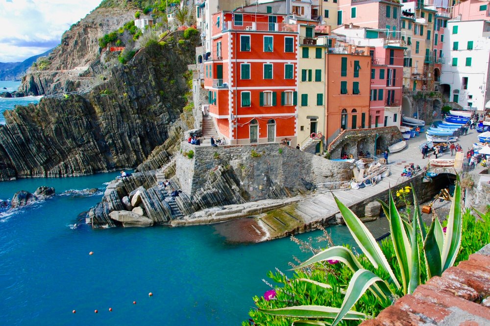 Bright buildings above a tiny harbor in Cinque Terre, with layered rock cliffs and deep blue sea beside them