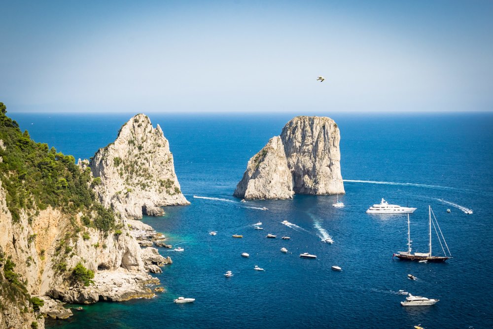 View of Capri’s Faraglioni rock formations offshore, with boats and yachts cruising through deep blue water under a clear sky