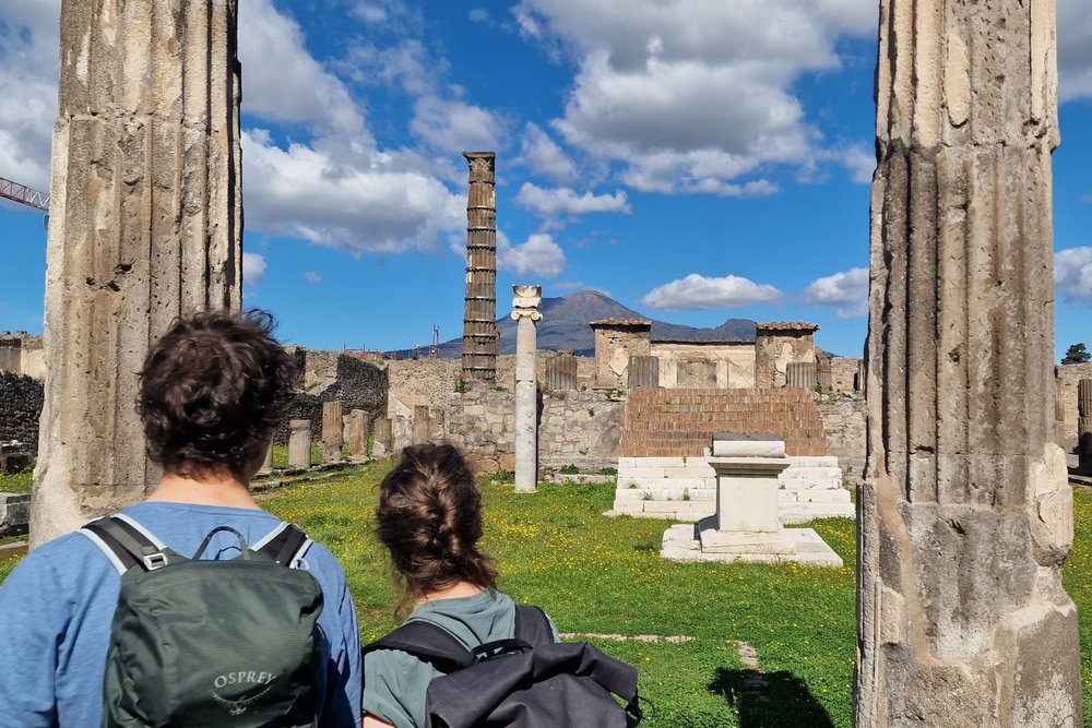 Two travelers with backpacks walk through Pompeii’s stone columns and ruins, with Mount Vesuvius under a blue sky
