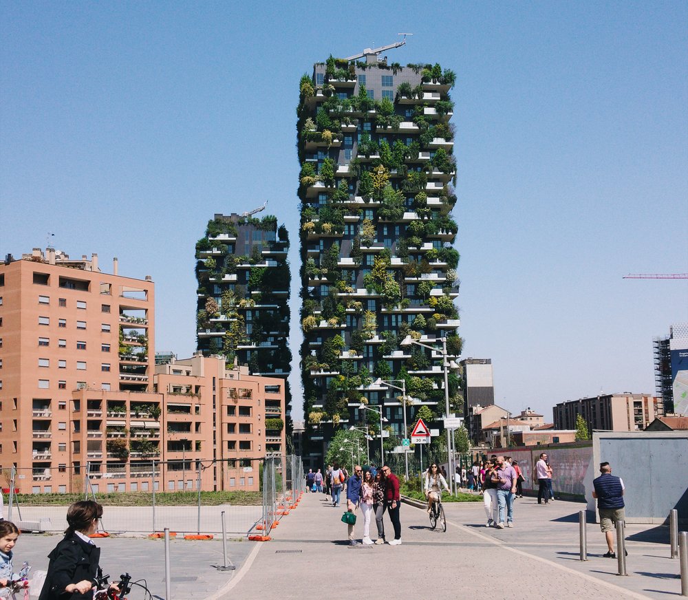 A street crowded with people walking and biking, with a salmon-colored building and vertical gardens in the background.