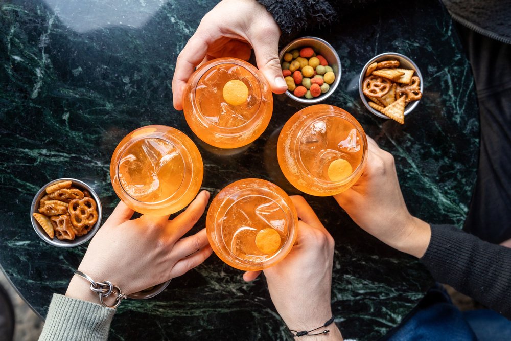 Four hands reaching for four cups filled with an orange liquid, surrounded by three small bowls of bar snacks.