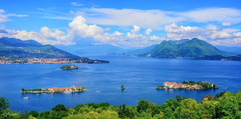 A body of water with trees in the foreground, centered parallel islands, and mountains and clouds in the background.