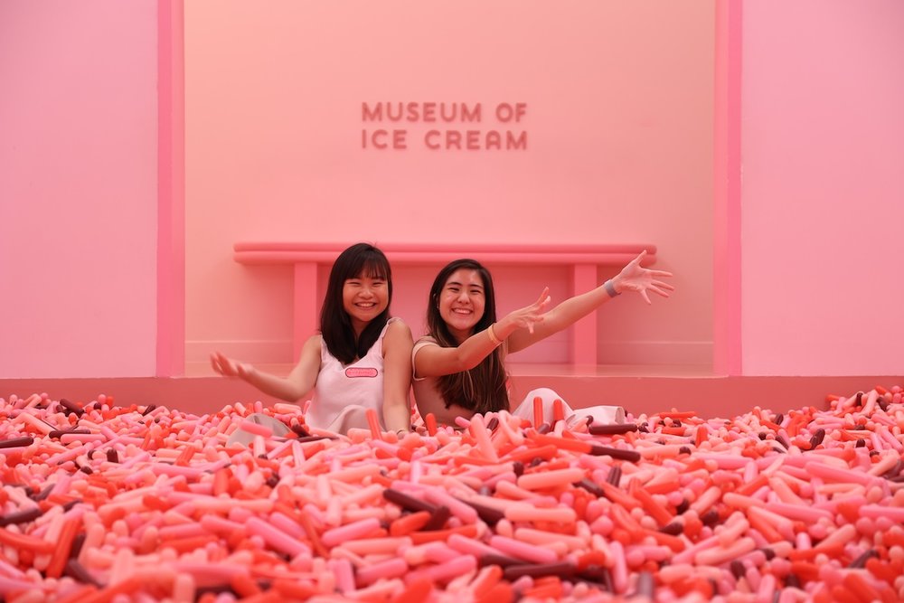 Two women smile and pose inside a pink sprinkle pit at the Museum of Ice Cream Singapore against a pink wall.