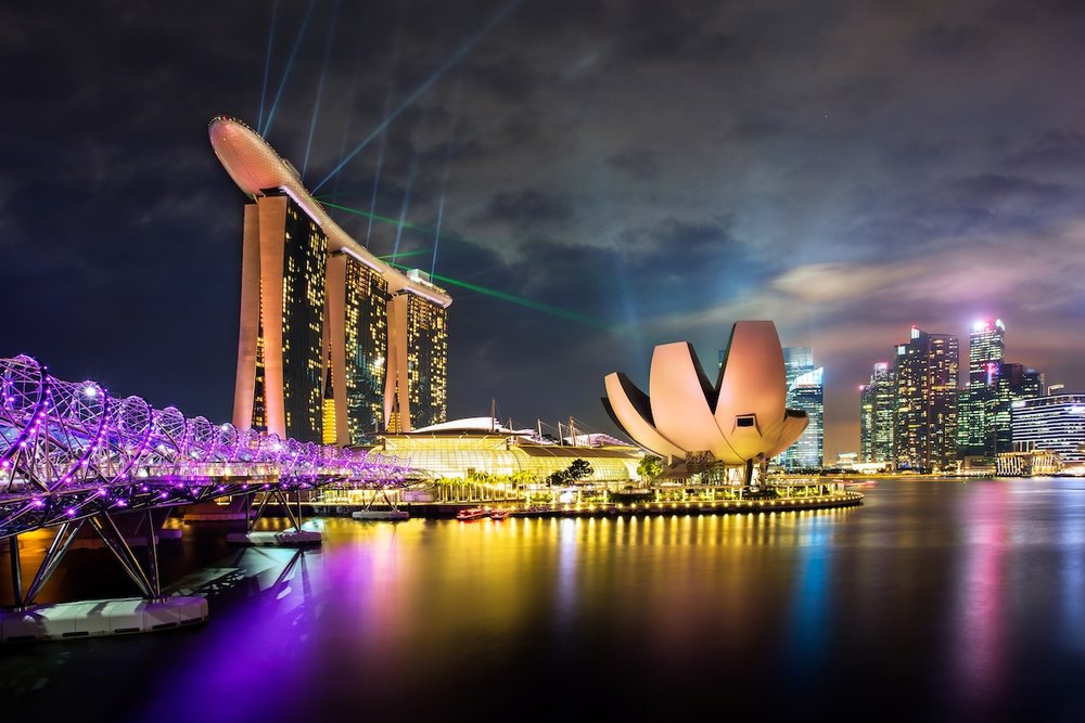 A nighttime cityscape of Marina Bay, Singapore, showing the purple-lit Helix Bridge leading toward the lotus-shaped ArtScience Museum and the Marina Bay Sands hotel.
