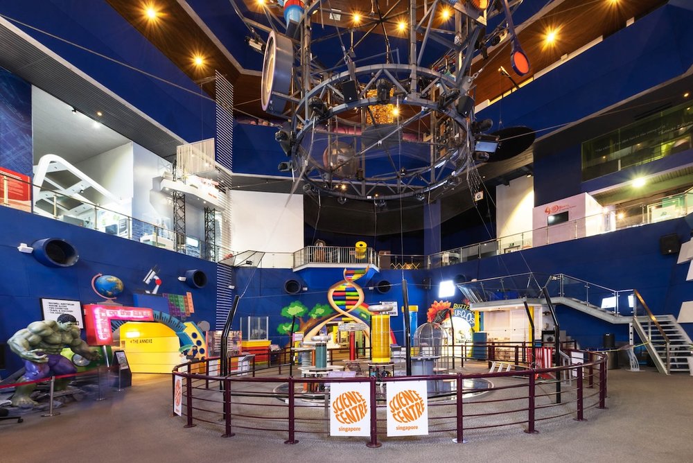 A wide-angle view of an indoor science museum with blue walls, multiple levels, and scientific displays. A large metal structure hangs from the ceiling over a fenced-off electrical exhibit.
