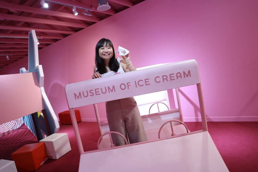 A woman smiling behind a pink stationary vehicle prop with "MUSEUM OF ICE CREAM" written on the front. The room has solid pink walls and a dark red floor.