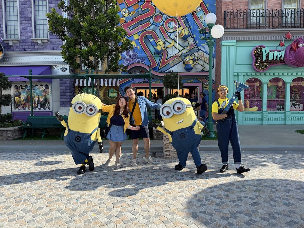 Two people stand between two large Minion mascots on a cobblestone street. To the right, a performer in a yellow shirt and overalls holds a blue megaphone prop against a backdrop of colorful USS buildings.