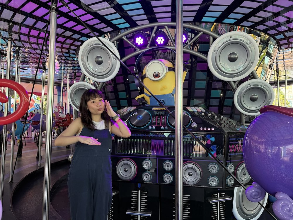 A woman stands beside a decorative DJ booth featuring a one-eyed Minion wearing headphones.