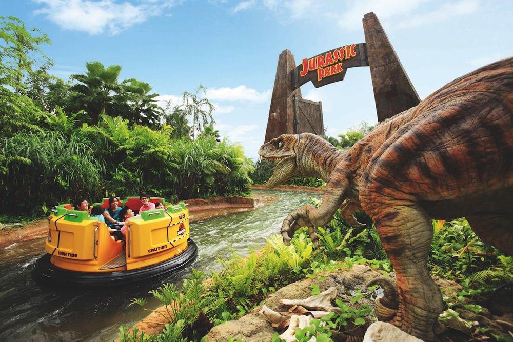 A yellow river rapids raft carrying several people floats down a water channel. In the foreground, a realistic animatronic dinosaur stands among rocks and greenery, with the stone Jurassic Park entrance gate in the background.
