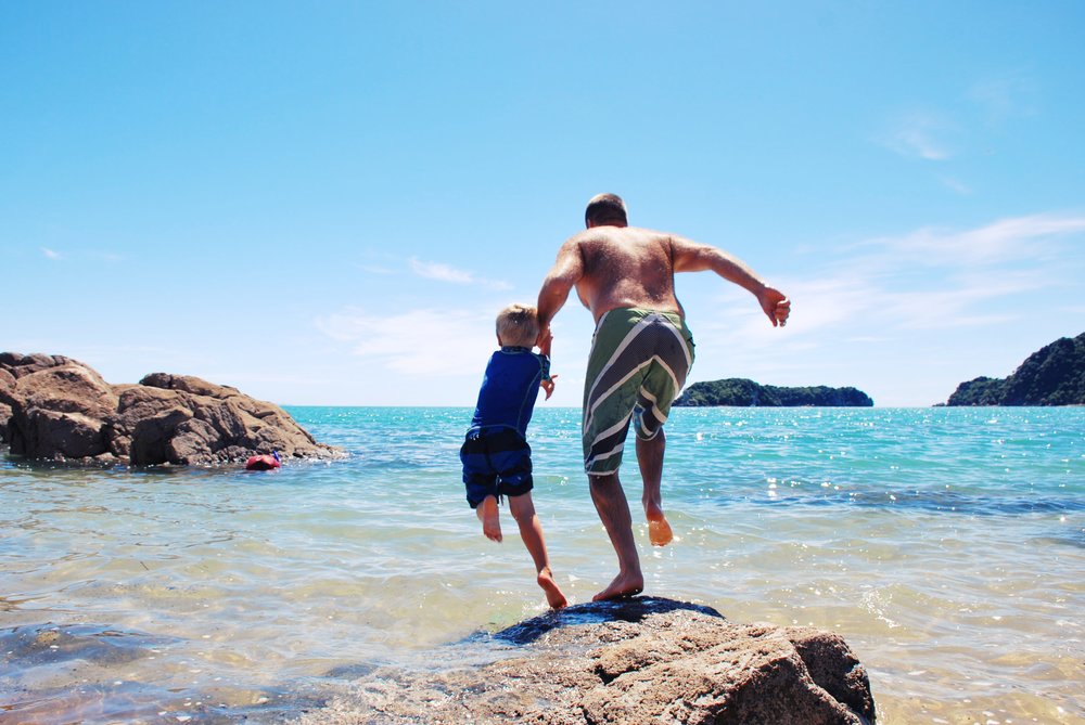 Father and son jumping into clear New Zealand beach water on a sunny day